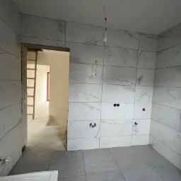 Łazienka w trakcie remontu: szare płytki podłogowe i białe marmurowe płytki ścienne, widoczne otwory na instalacje, drabina w przejściu do innego pomieszczenia, surowa żarówka zwisająca z sufitu.