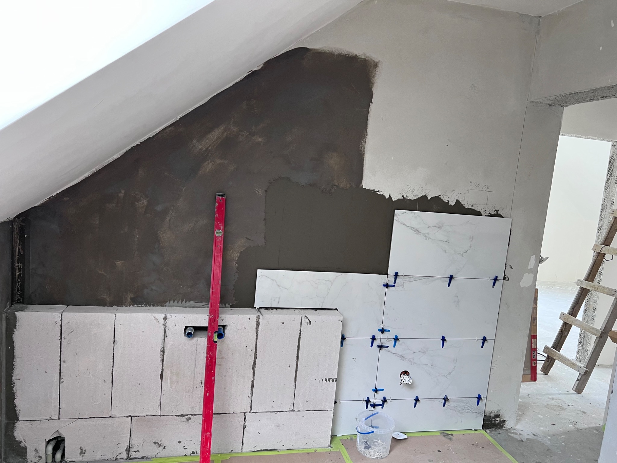 Montaż płytek imitujących marmur w łazience na poddaszu, widoczne nieotynkowane ściany, poziomica i wiadro z krzyżykami dystansowymi.