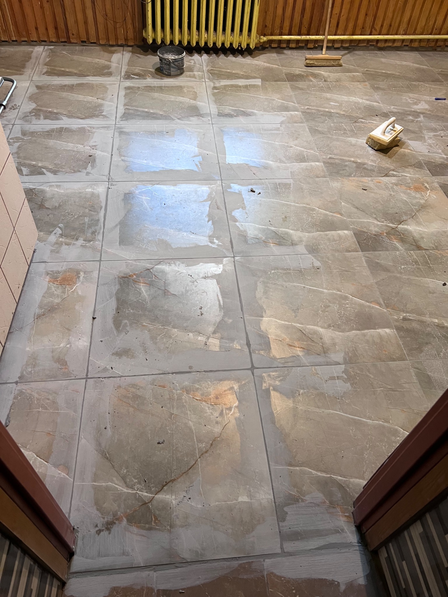 Szare płytki podłogowe imitujące marmur, z widocznymi śladami świeżo nałożonej fugi, narzędzia glazurnicze w tle, fragment grzejnika żeberkowego i drewnianej boazerii.