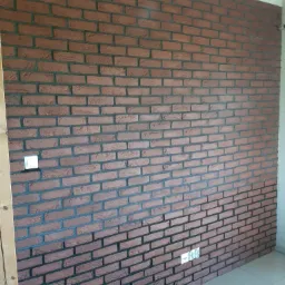 Ściana wykończona brązową cegłą klinkierową z ciemną fugą, widoczny włącznik światła i gniazdka elektryczne.