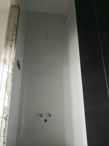 Surowa ściana łazienki z widocznymi instalacjami wodnymi i elektrycznymi, przygotowana do dalszych prac wykończeniowych, z fragmentem surowego muru po lewej stronie i czarną płytą po prawej.