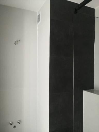 Surowa łazienka w trakcie wykańczania: białe ściany, czarne płytki w kabinie prysznicowej, widoczne przyłącza wodne i wentylacja.