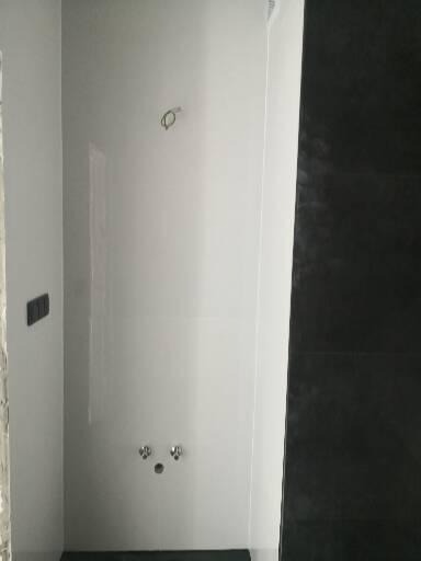 Świeżo wykończona, minimalistyczna łazienka z białymi płytkami ściennymi i czarną ścianą prysznicową, widoczne przyłącza wodne i niedokończone gniazdko elektryczne.