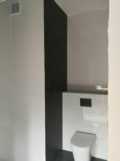 Nowoczesna łazienka z białą toaletą podwieszaną i czarną ścianą z płytek, widoczna kratka wentylacyjna na suficie.