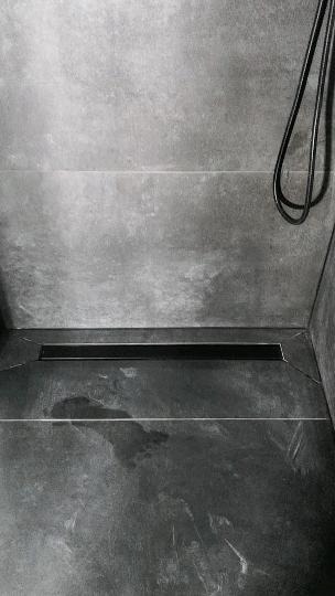 Nowoczesny odpływ liniowy w posadzce kabiny prysznicowej wyłożonej szarymi płytkami imitującymi kamień, z widocznym przewodem słuchawki prysznicowej.