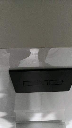 Czarny panel sterujący spłuczką wbudowaną w ścianę, zamontowany na białych, błyszczących płytkach w łazience.