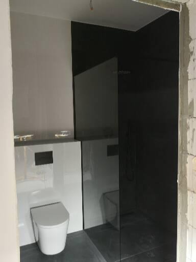 Nowoczesna łazienka w trakcie wykańczania: biała toaleta wisząca, czarna kabina prysznicowa typu walk-in z widocznym odpływem liniowym, białe płytki ścienne, szara podłoga, surowy beton na krawędzi...