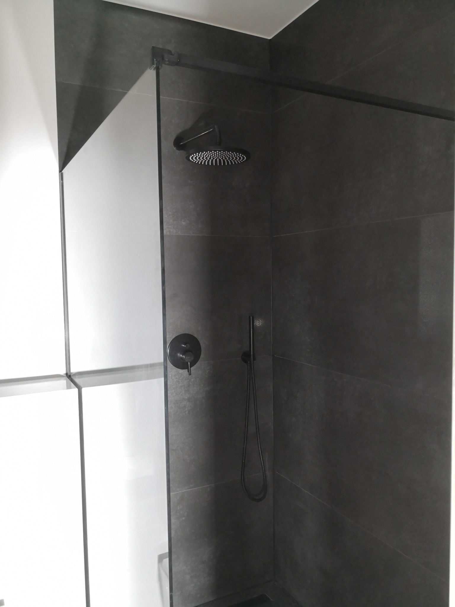 Nowoczesna kabina prysznicowa z ciemnoszarymi płytkami, deszczownicą i czarną armaturą, widok z boku, z fragmentem białych szafek w tle.