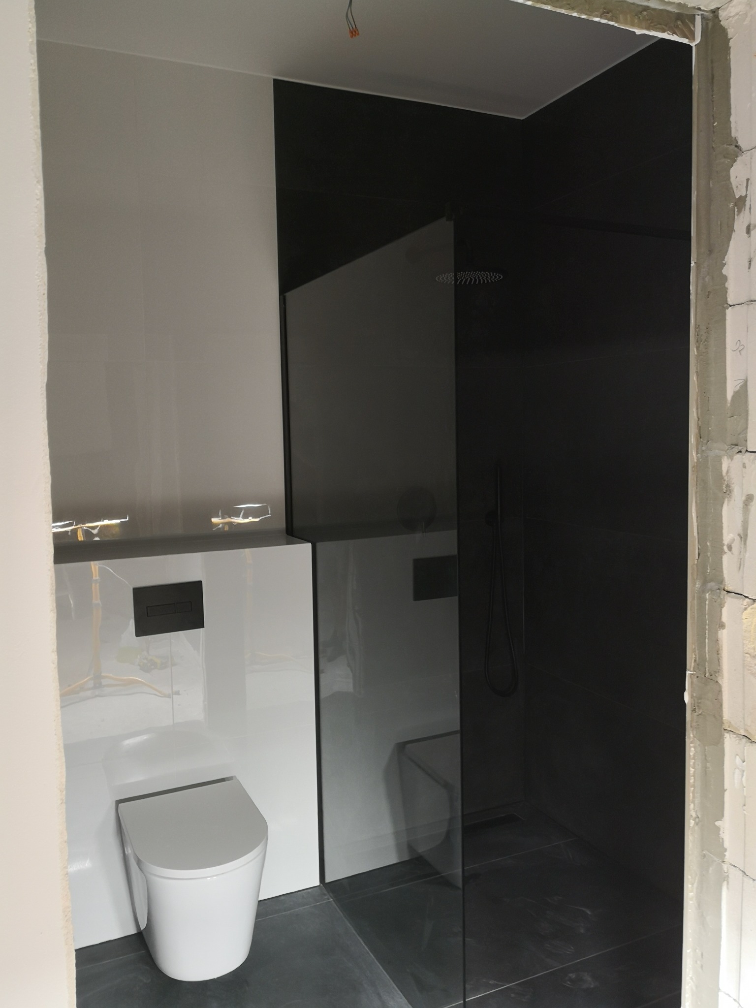 Nowoczesna łazienka w trakcie wykończenia, z kabiną prysznicową typu walk-in z czarnymi płytkami i podwieszaną toaletą w białej obudowie, widoczne niedokończone ściany.