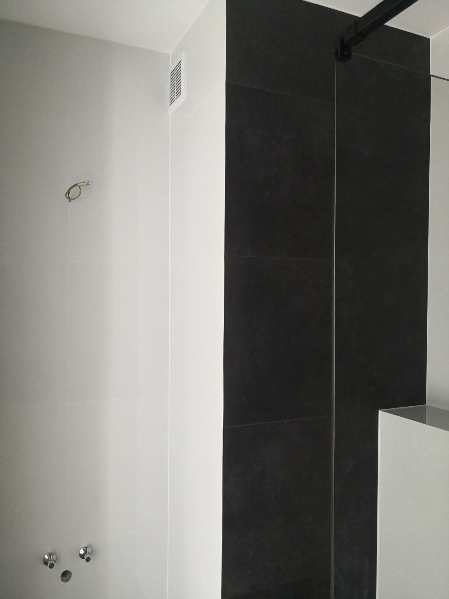 Wnętrze łazienki w trakcie wykańczania: białe płytki na ścianach z widocznymi przyłączami hydraulicznymi, czarne płytki w strefie prysznicowej, wentylacja mechaniczna, surowy kabel elektryczny.