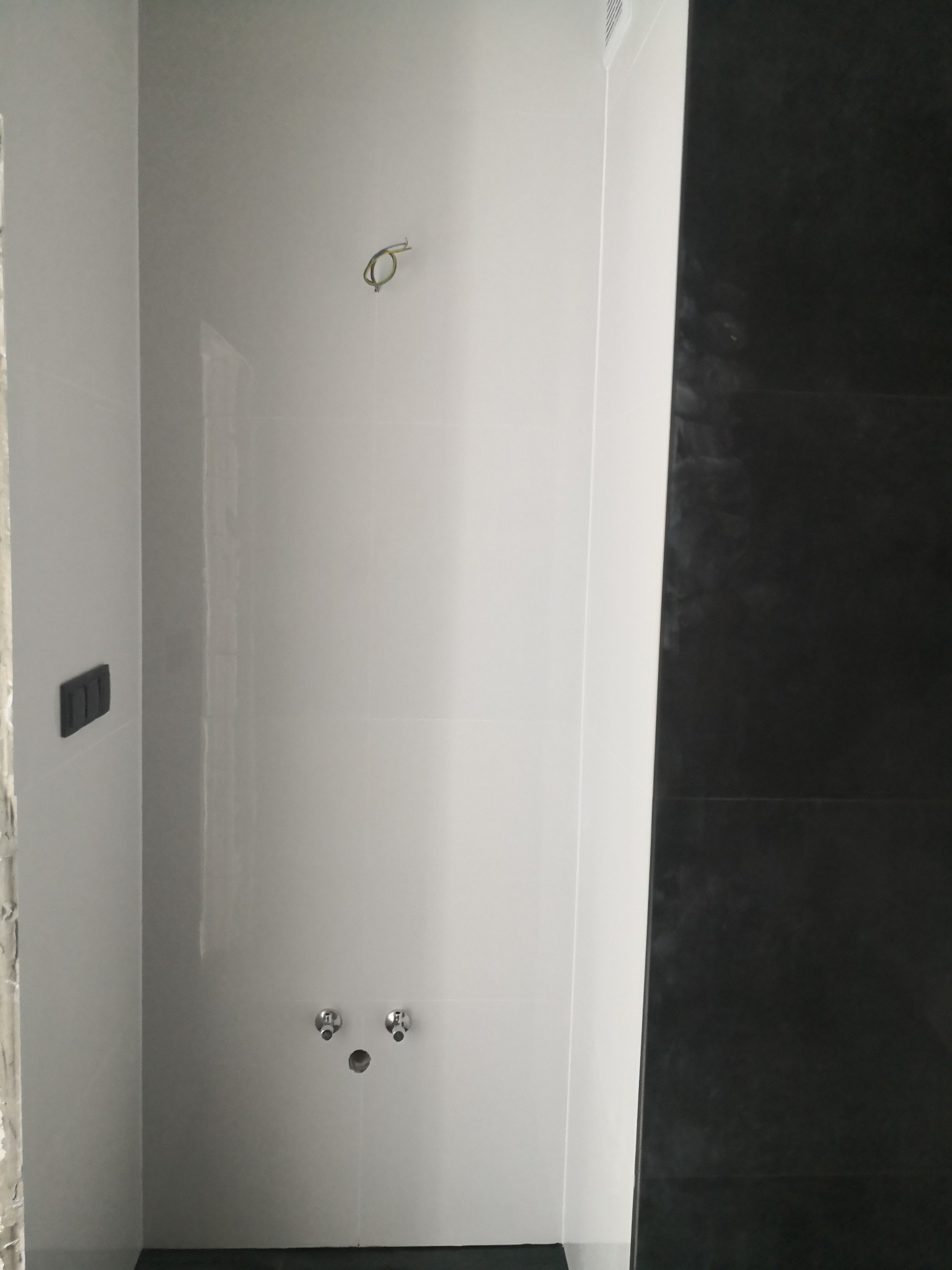 Wnętrze kabiny prysznicowej w trakcie wykańczania: białe płytki na ścianach z wyprowadzonymi przyłączami wodnymi i niedokończonymi fugami, obok ściana wyłożona czarnymi płytkami.