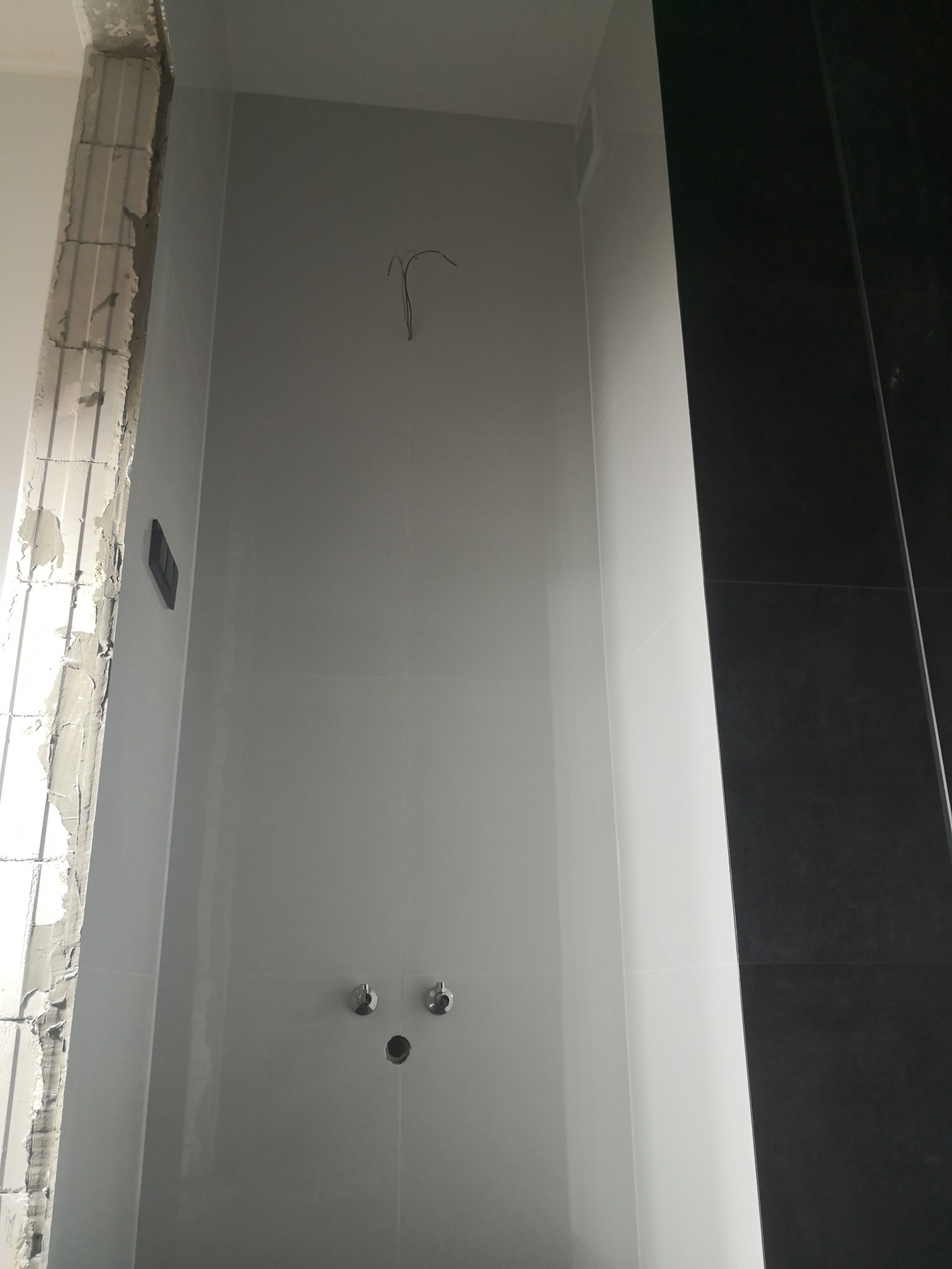 Łazienka w trakcie wykończenia: białe płytki na ścianach, widoczne przyłącza wody, niedokończona ściana z lewej strony, czarne płytki z prawej strony.