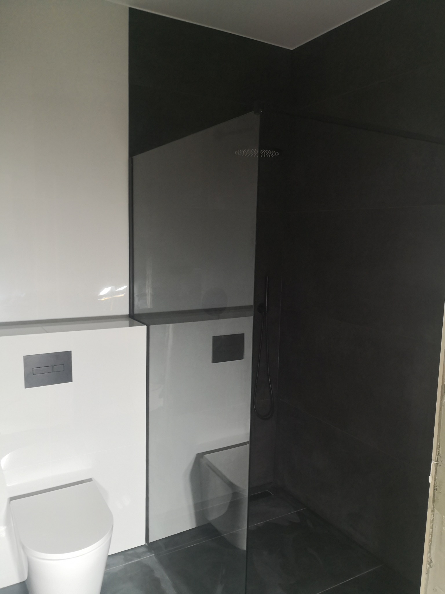 Nowoczesna łazienka w czarno-białej kolorystyce z kabiną prysznicową typu walk-in i podwieszaną toaletą, widoczne szare płytki podłogowe i minimalistyczna armatura.