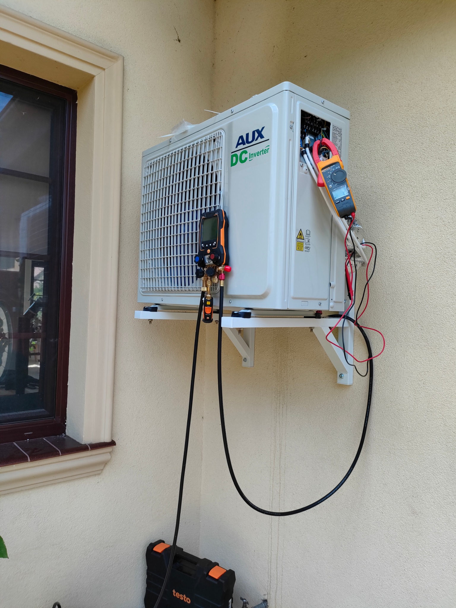 Biała jednostka zewnętrzna klimatyzacji AUX DC Inverter zamontowana na ścianie budynku, podłączona do mierników kontrolnych podczas serwisu, widoczna skrzynka narzędziowa Testo.