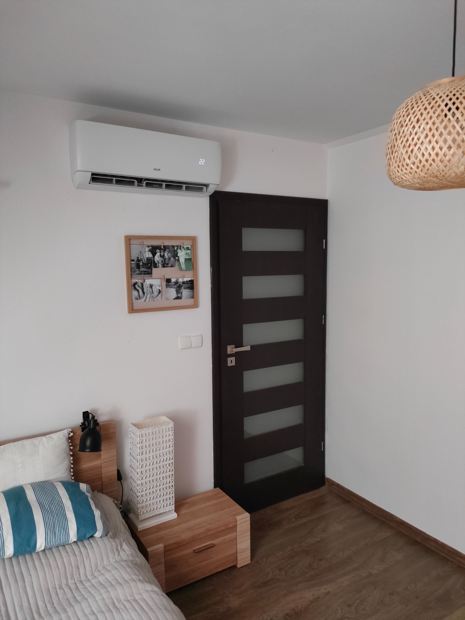 Biały klimatyzator AUX zamontowany na ścianie w sypialni nad łóżkiem, widoczny również drewniany stolik nocny z lampką i dekoracyjną lampą, drewniana podłoga i drzwi z przeszkleniami.