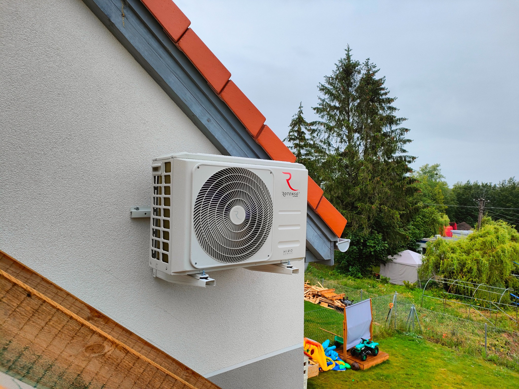 Zewnętrzna jednostka klimatyzacji Rotenso zamontowana na elewacji domu, widok na ogród z zabawkami.