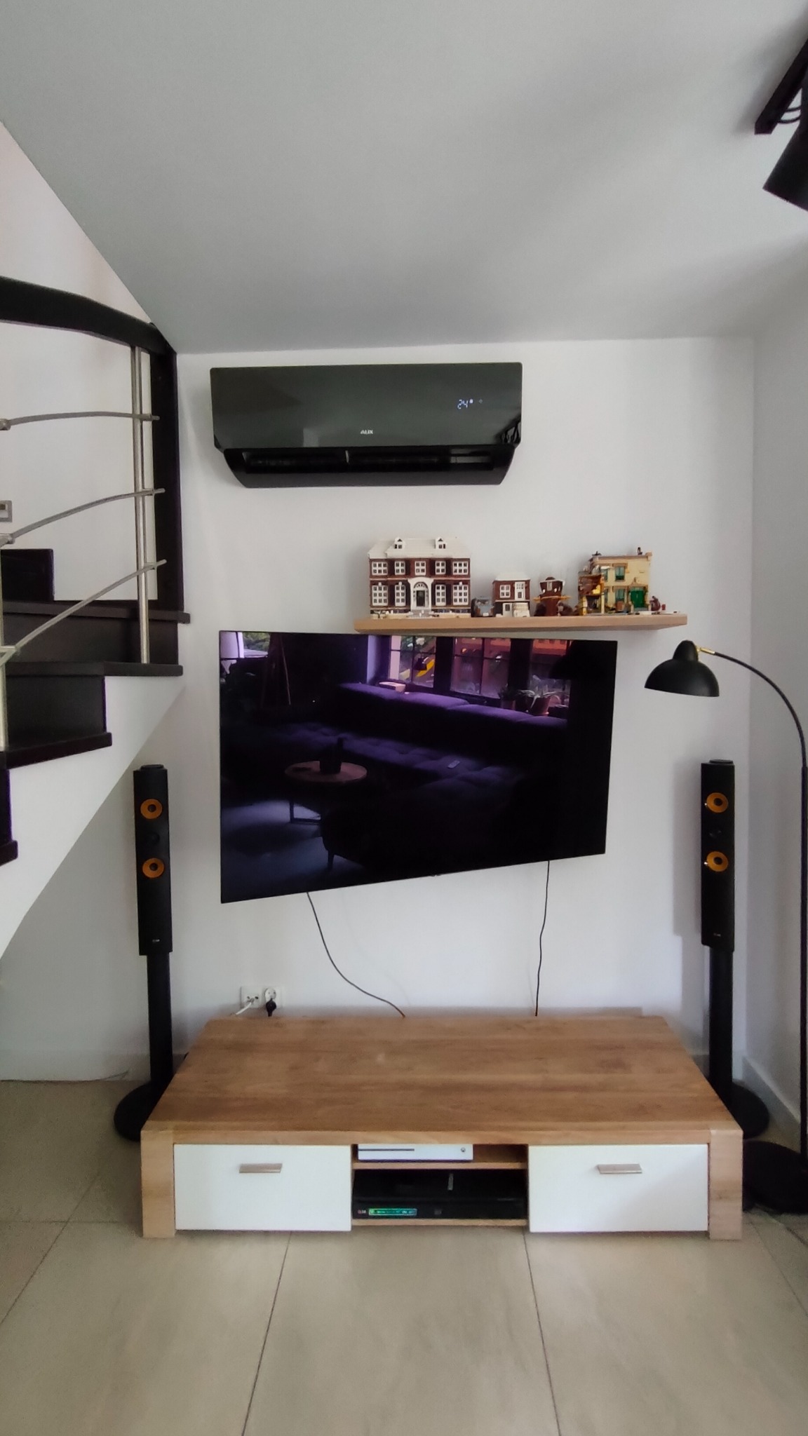 Czarny klimatyzator ścienny marki AUX zamontowany w nowoczesnym salonie, widoczny nad telewizorem i półką z modelami budynków LEGO.