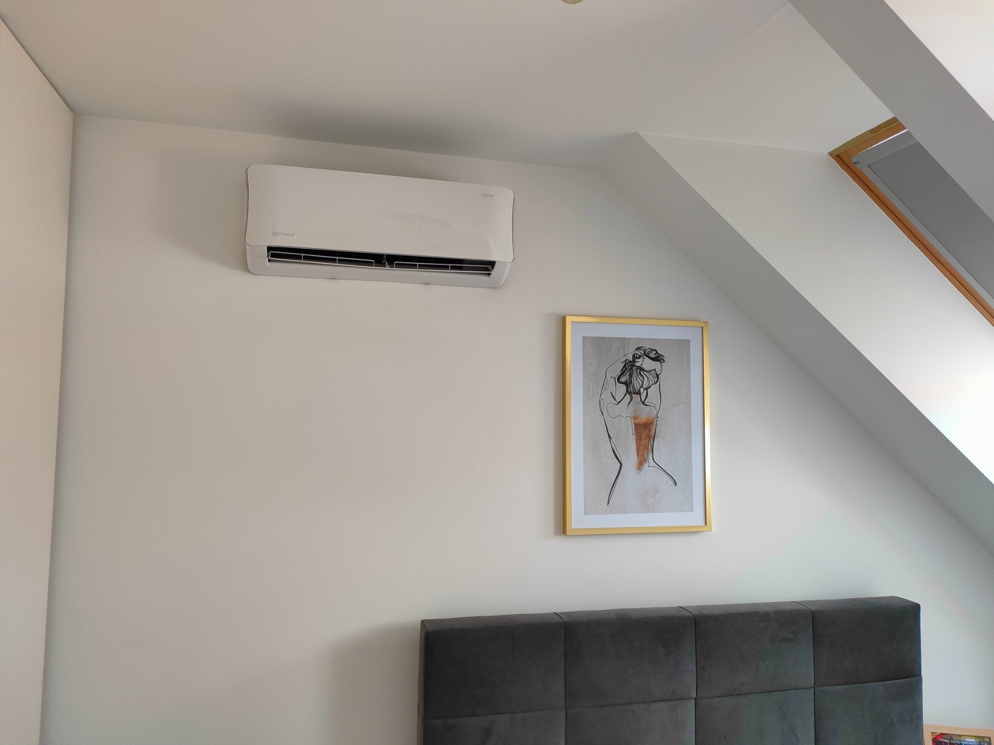 Biała jednostka klimatyzacyjna Rotenso zamontowana na białej ścianie w pokoju na poddaszu, obok rama okienna i obrazek w złotej ramie nad wezgłowiem łóżka.