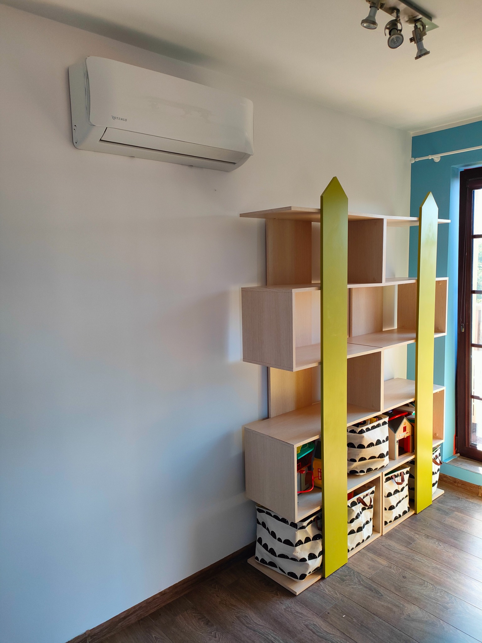 Biała klimatyzacja Rotenso na ścianie w pokoju dziecięcym, obok regał z zabawkami i koszami do przechowywania.