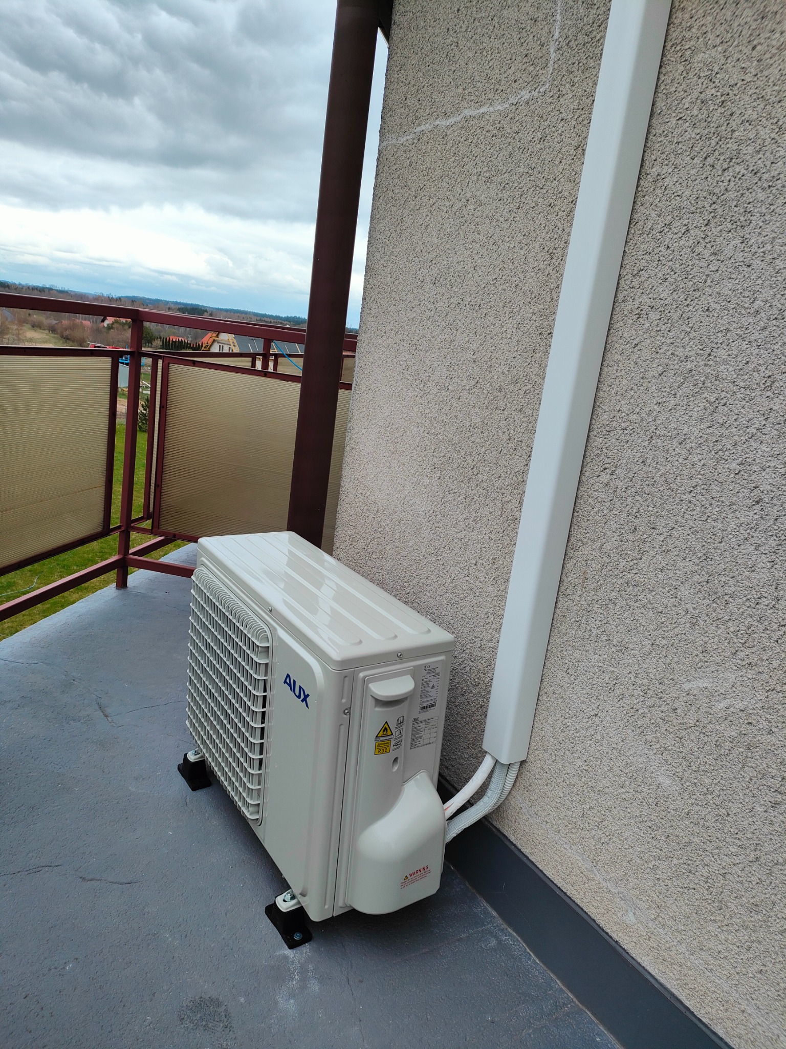 Zewnętrzna jednostka klimatyzacji AUX zamontowana na balkonie domu jednorodzinnego, z widoczną maskownicą przewodów na elewacji. W tle krajobraz z zabudową mieszkalną.
