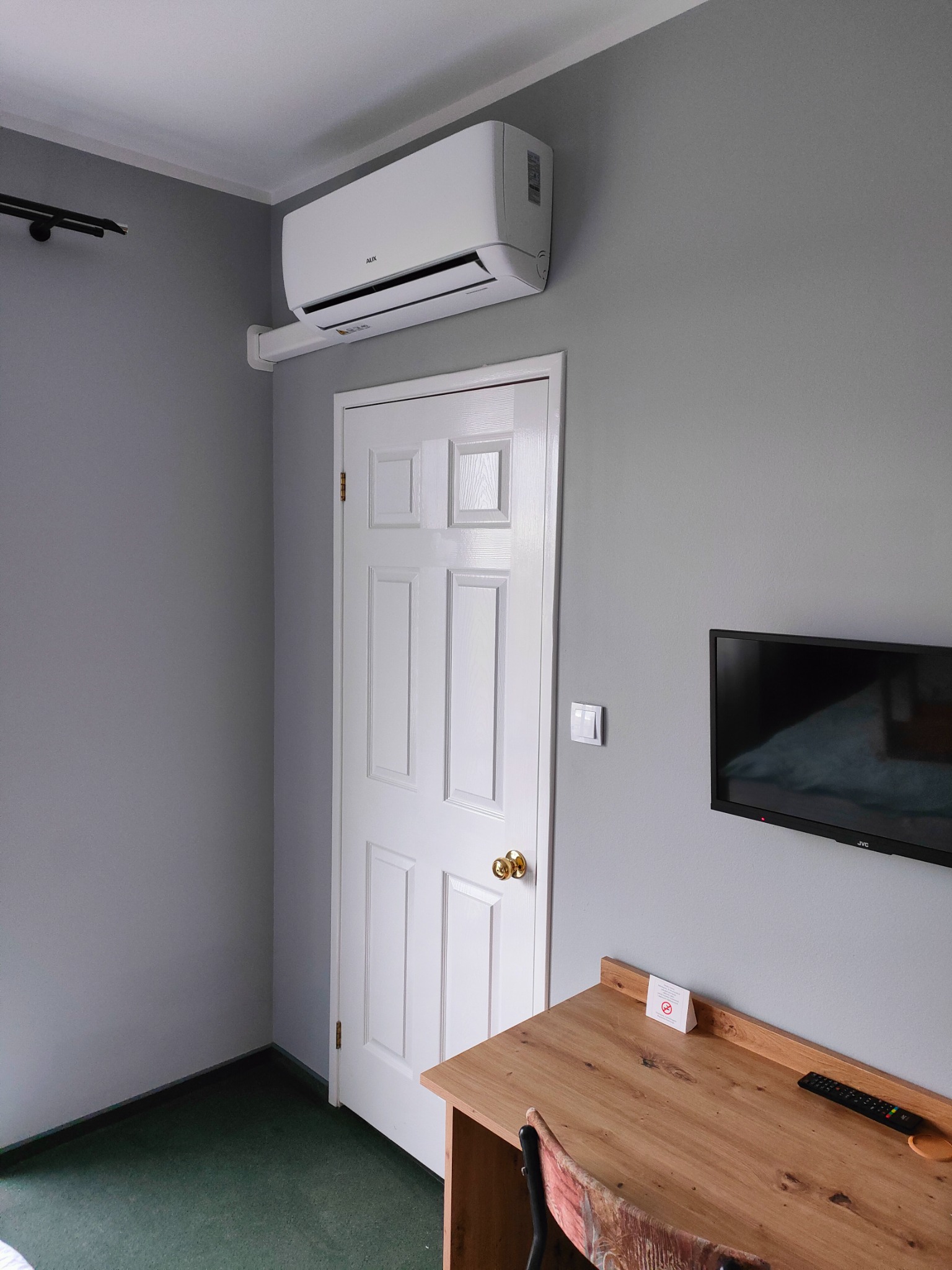 Biały klimatyzator AUX zamontowany nad białą, panelową ścianą drzwi w pokoju z szarą ścianą i biurkiem z drewna.