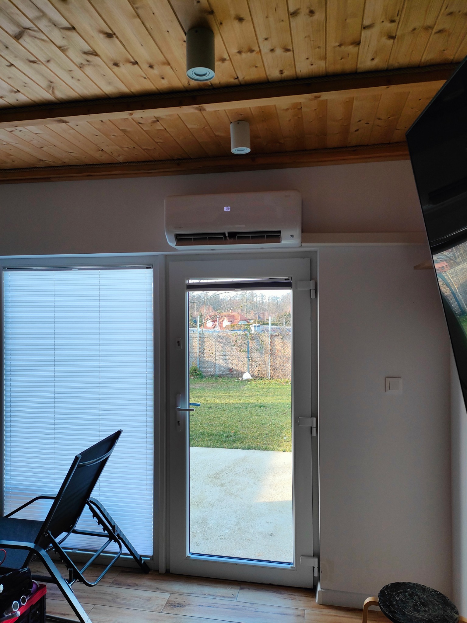 Klimatyzator ścienny zamontowany nad drzwiami balkonowymi w pomieszczeniu z drewnianym sufitem i widokiem na ogród.