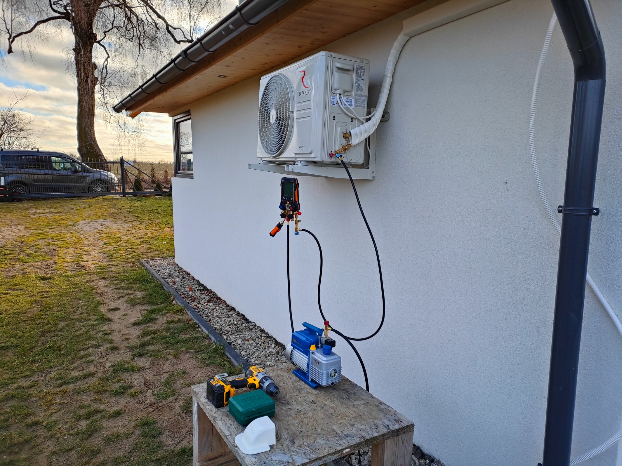 Biała jednostka zewnętrzna klimatyzatora zamontowana na ścianie domu, podłączona do pompy próżniowej i manometru, na drewnianym stoliku obok wiertarka akumulatorowa.