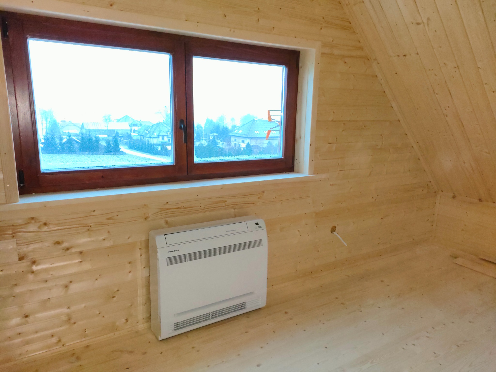 Biały klimatyzator podokienny marki Innova w drewnianym pokoju na poddaszu, widok na zewnątrz przez okno.