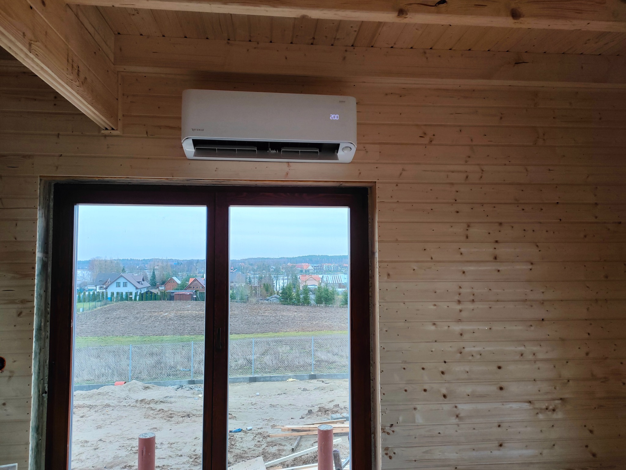 Biały klimatyzator marki Rotenso zamontowany nad oknem w drewnianym domu w trakcie budowy, widok na zewnątrz.