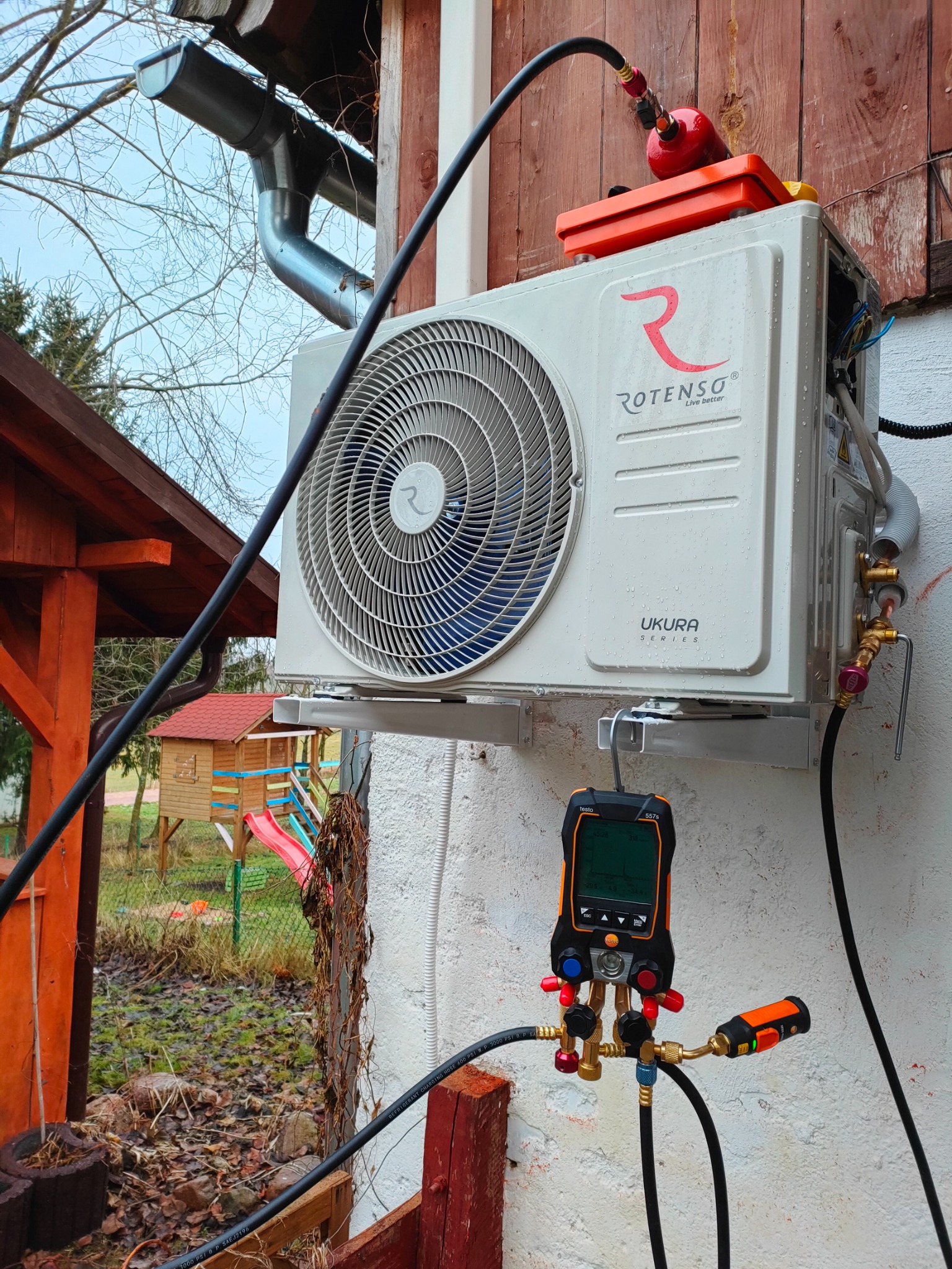 Zewnętrzna jednostka klimatyzacji Rotenso Ukura Series zamontowana na ścianie budynku, podłączona do manometru cyfrowego Testo 557s i butli z czynnikiem chłodniczym, na tle ogrodu z domkiem...