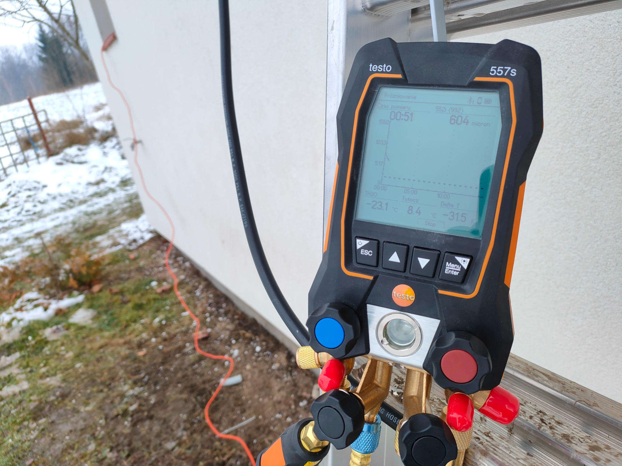 Miernik cyfrowy Testo 557s podłączony do instalacji klimatyzacyjnej, wskazujący wartość próżni w mikronach, ustawiony na zewnątrz budynku w trakcie serwisu.