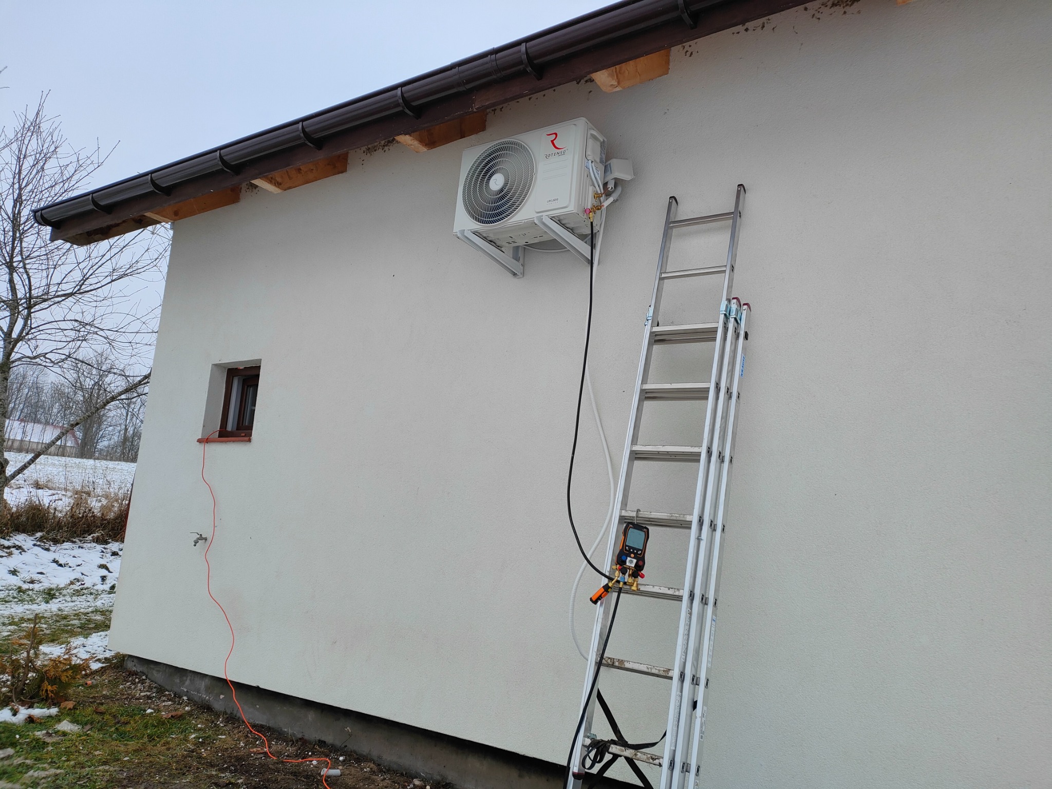 Zewnętrzna jednostka klimatyzacji Rotenso zamontowana na ścianie domu, widoczna drabina oparta o ścianę z podłączonym miernikiem ciśnienia czynnika chłodniczego.