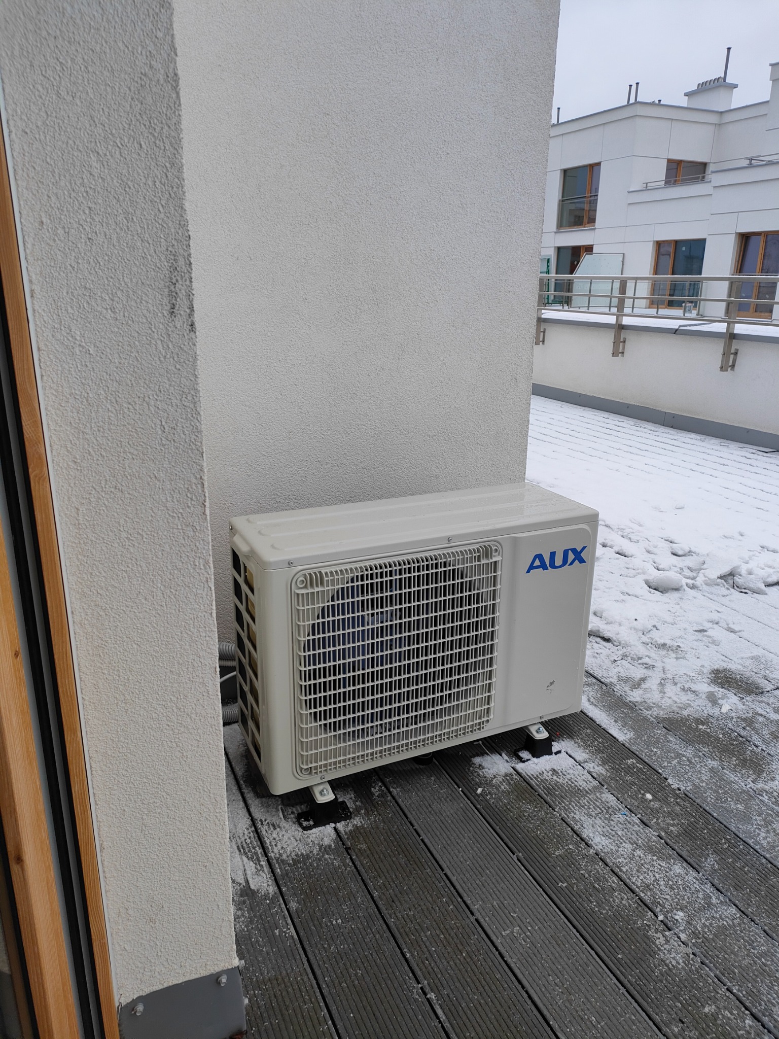 Zewnętrzna jednostka klimatyzacji AUX zamontowana na drewnianym tarasie w zimowej scenerii, z widocznym śniegiem na podłodze i w tle zabudowania wielorodzinnego.