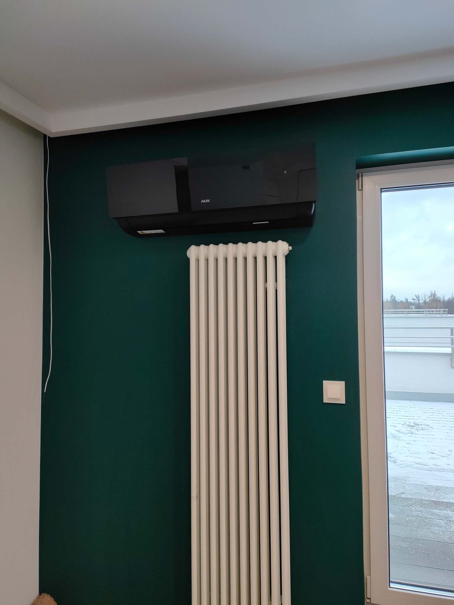 Czarny klimatyzator ścienny marki AUX zamontowany w pokoju na zielonej ścianie nad białym grzejnikiem żeberkowym, widok z okna na zewnątrz.
