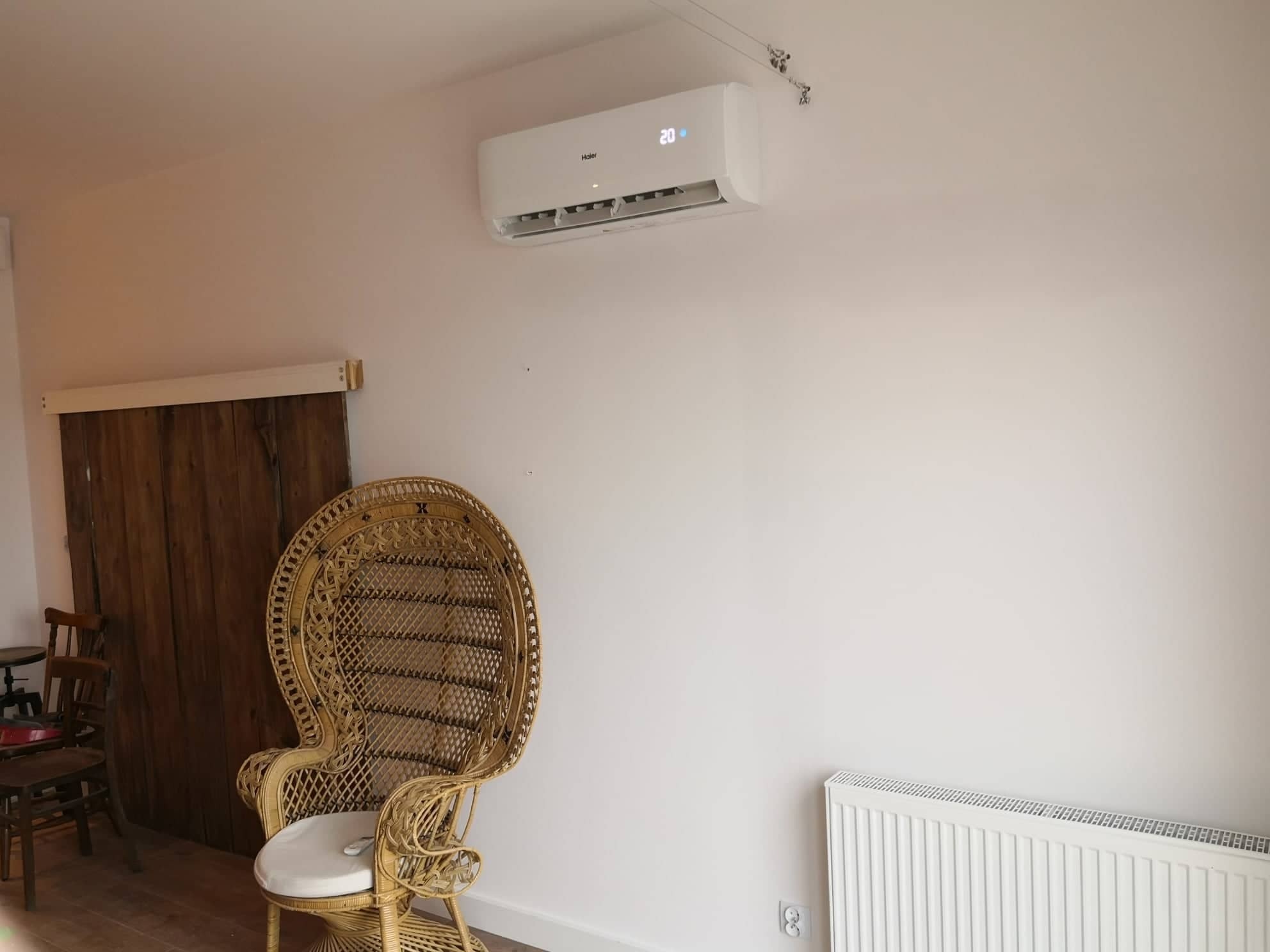 Biały klimatyzator Haier zamontowany na ścianie w jasnym pokoju z rattanowym fotelem pawim i drewnianymi drzwiami przesuwnymi w tle.