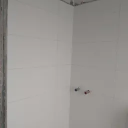 Białe płytki na ścianie w rogu pomieszczenia, widoczne przyłącza wodne z niebieską i czerwoną zaślepką, oraz metalowa listwa w rogu.