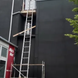 Elewacja budynku pomalowana na czarno, z rusztowaniem i drabiną ustawioną do okna na wyższej kondygnacji. W tle widoczny fragment budynku z czerwonym napisem 'Mila's Obst'.