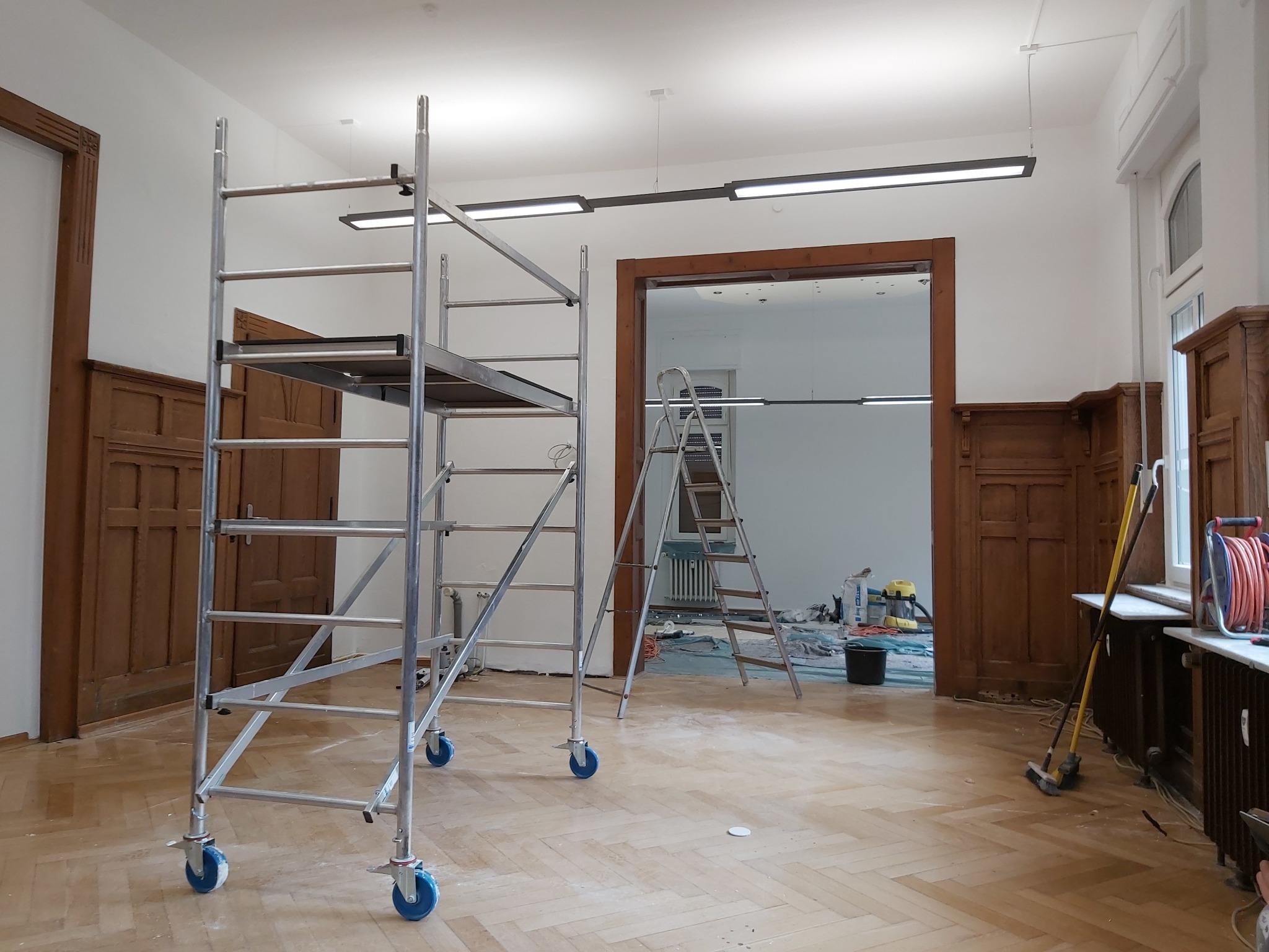 Wnętrze w trakcie remontu: aluminiowe rusztowanie na kółkach, drabina, widoczne ślady prac malarskich na podłodze parkietowej, ściany pomalowane na biało, oryginalna stolarka drzwiowa w kolorze...