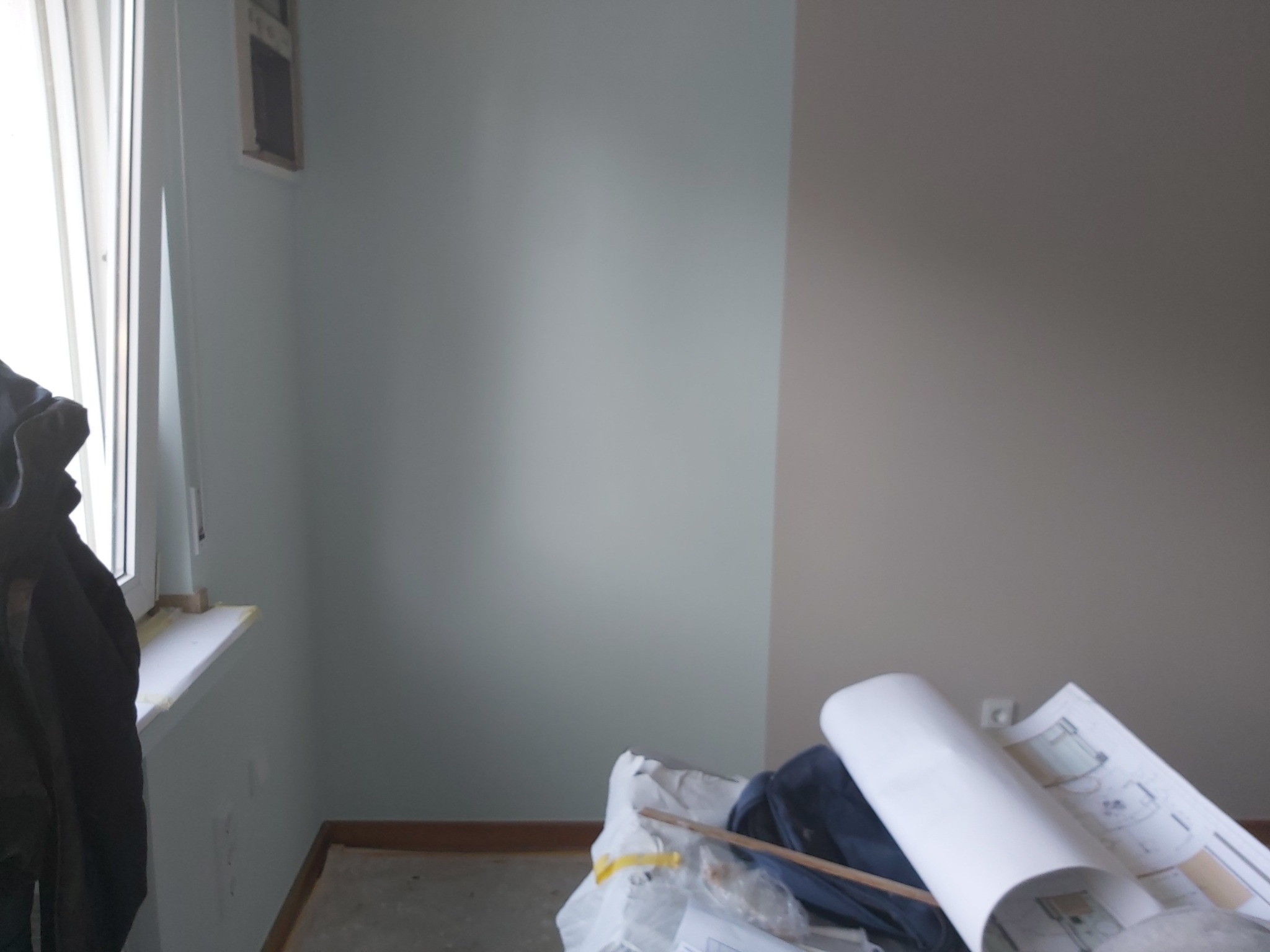 Świeżo pomalowane wnętrze z widocznym przejściem kolorów na ścianie, z pozostawionymi planami i narzędziami malarskimi na podłodze.