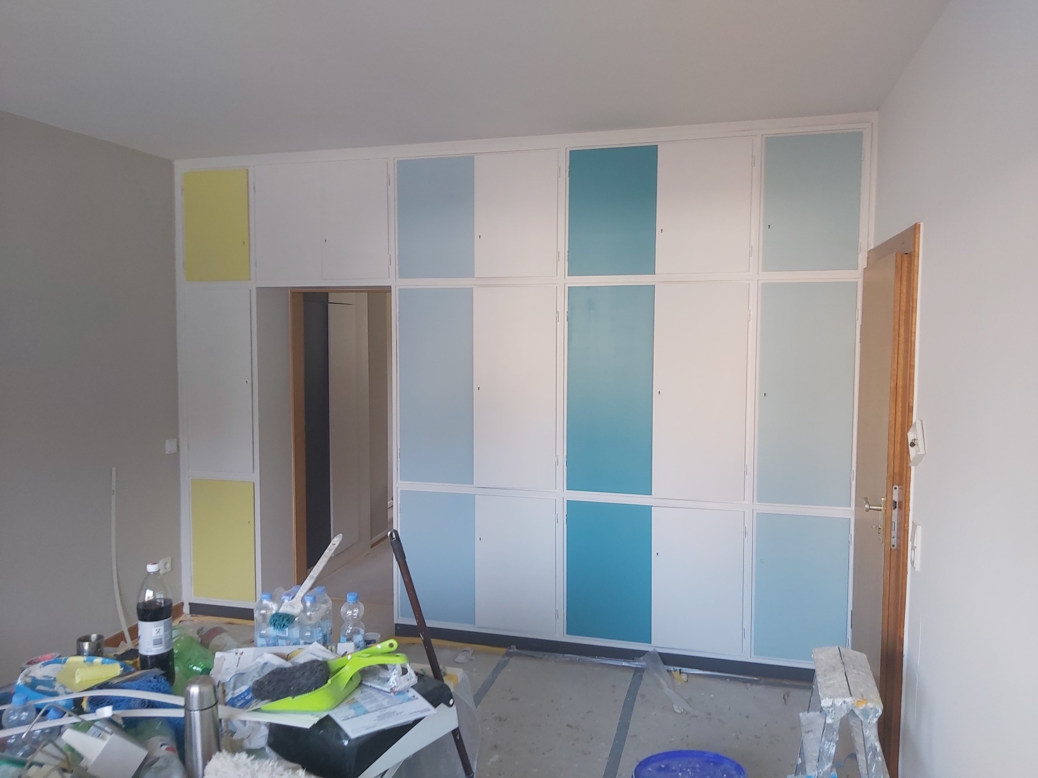 Wnętrze pokoju w trakcie malowania: ściana z wbudowanymi szafkami z pomalowanymi na biało, błękitno i turkusowo frontami, zabezpieczona podłoga, widoczny bałagan i narzędzia malarskie.