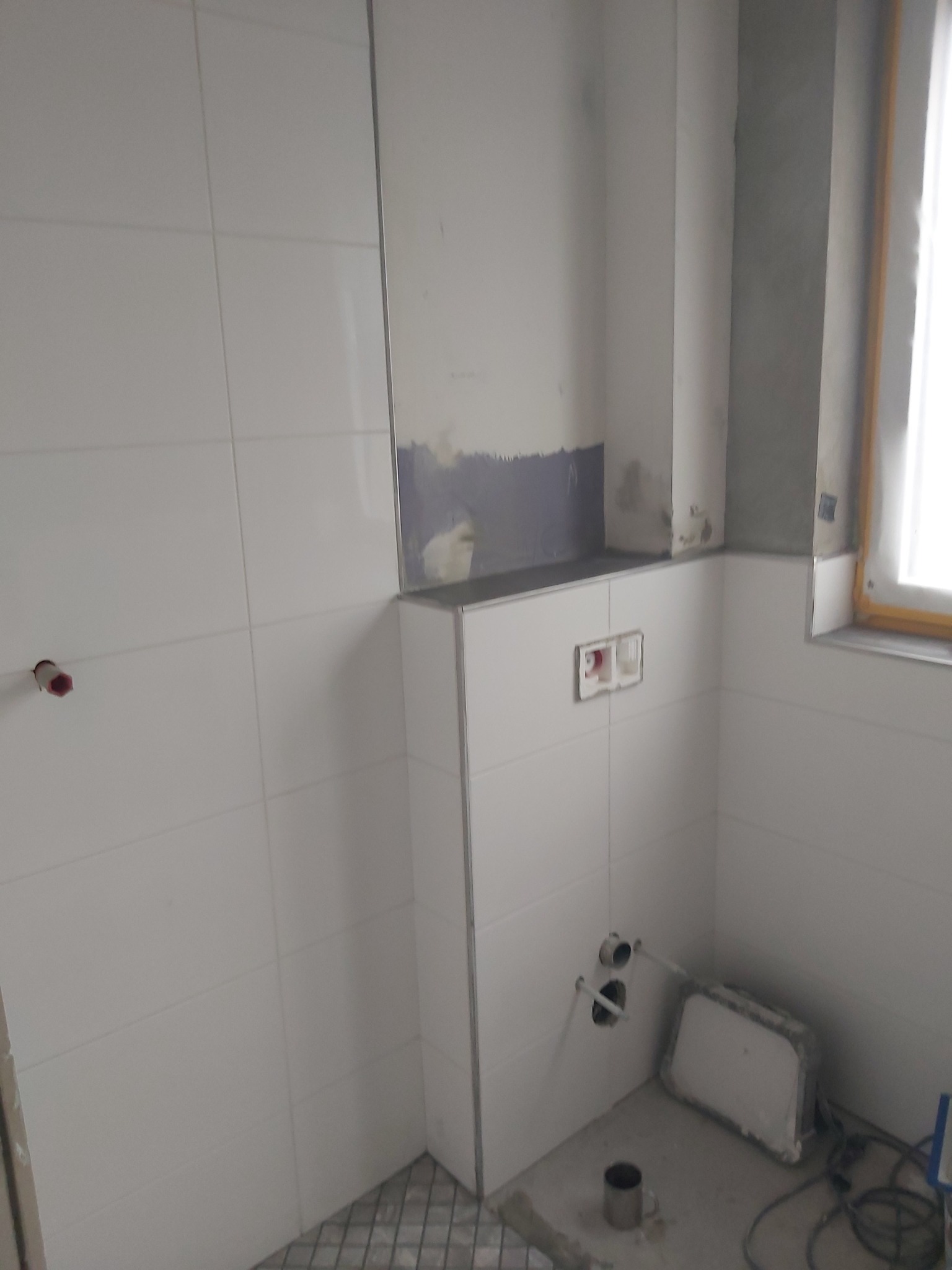 Wnętrze łazienki w trakcie wykańczania: białe, prostokątne płytki ceramiczne na ścianach, widoczne instalacje wodno-kanalizacyjne, fragment podłogi z mozaiką, niezabezpieczone kable elektryczne...