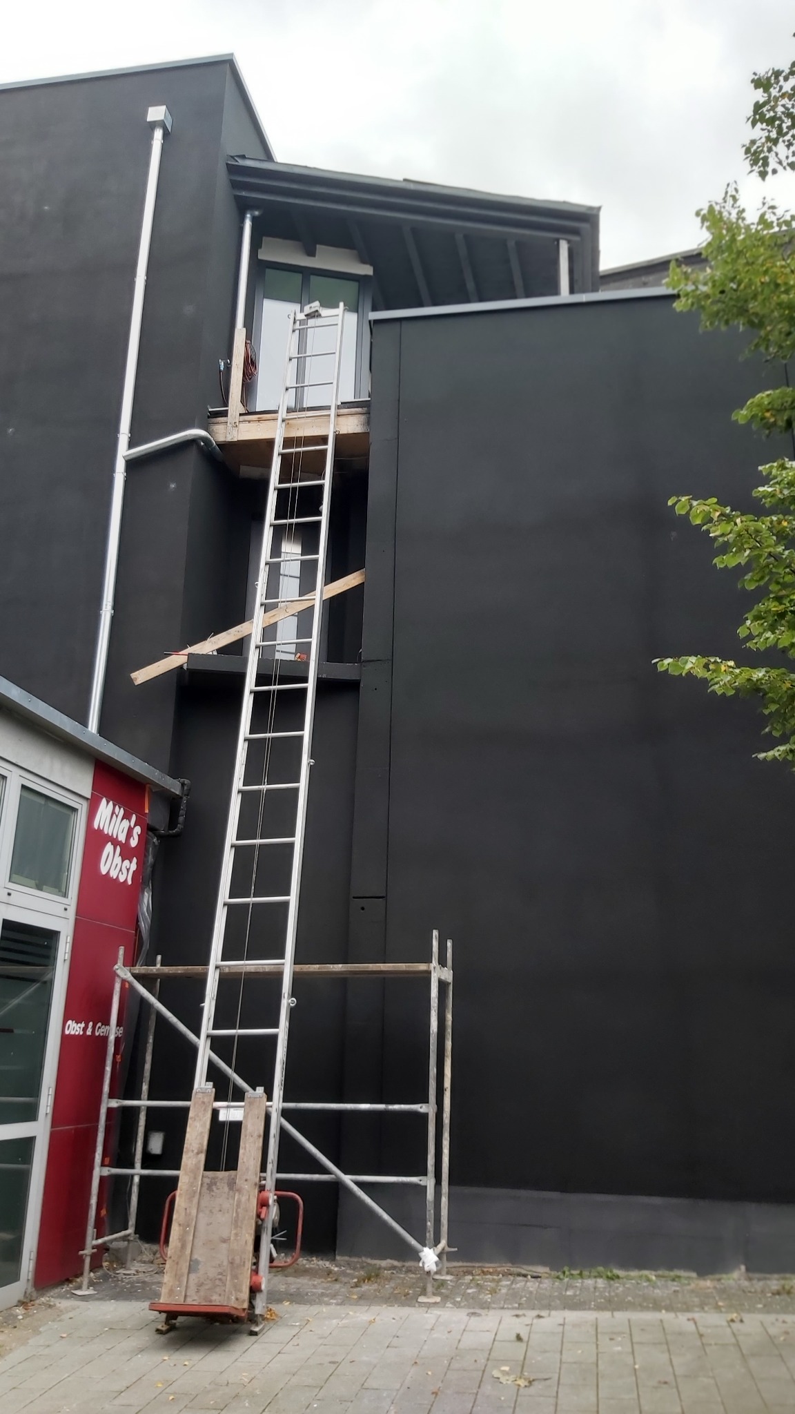 Elewacja budynku pomalowana na czarno, z rusztowaniem i drabiną ustawioną do okna na wyższej kondygnacji. W tle widoczny fragment budynku z czerwonym napisem 'Mila's Obst'.