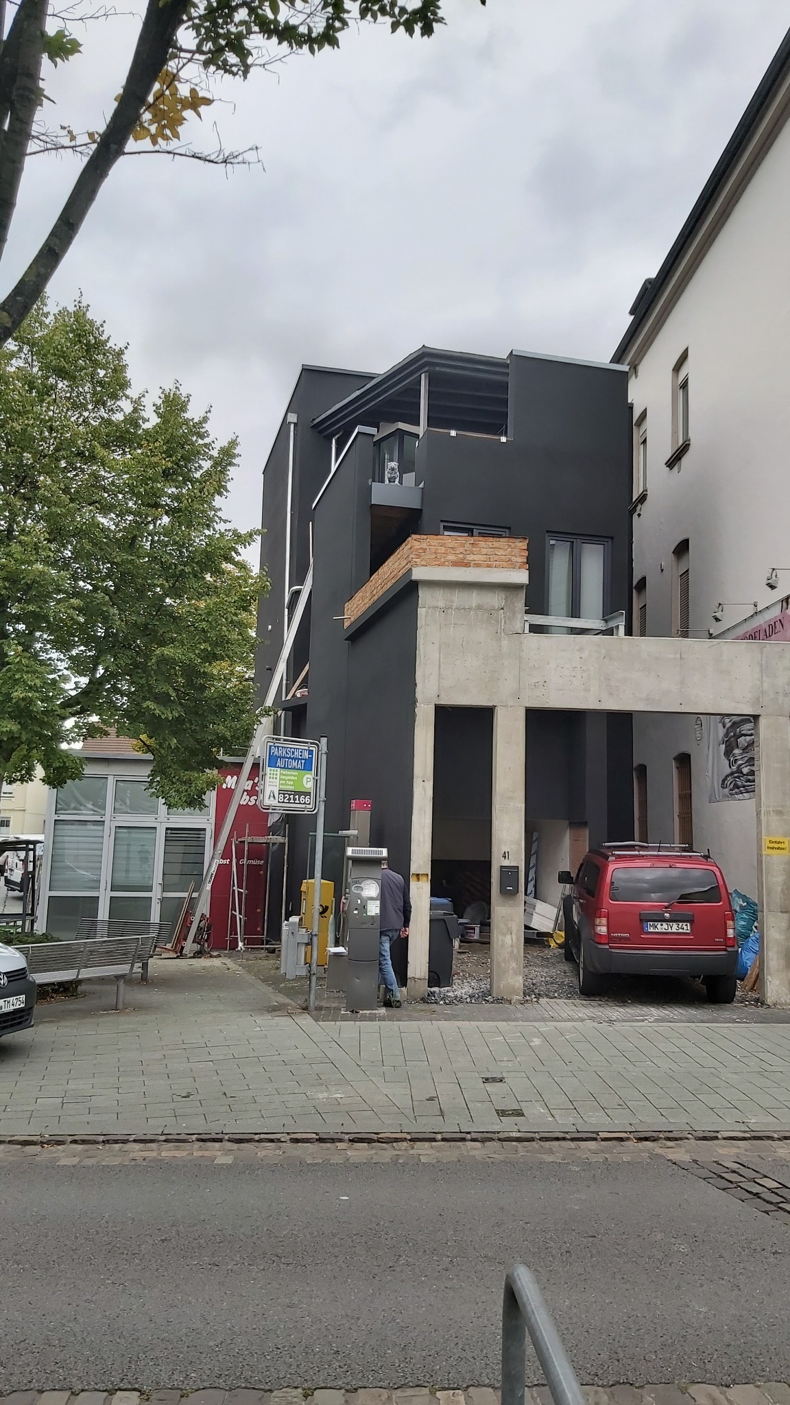 Czarny, nowocześnie otynkowany budynek z odsłoniętymi elementami konstrukcyjnymi z betonu, widoczny z chodnika obok parkomatu i zaparkowanego czerwonego samochodu.