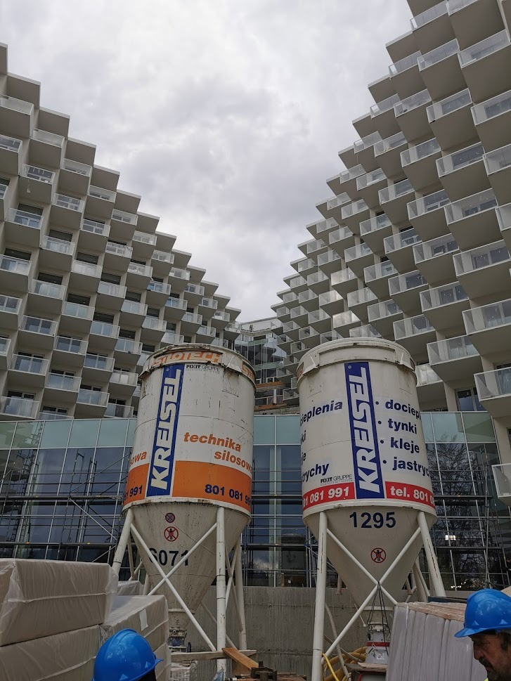 Dwa silosy budowlane marki Kreisel na tle nowoczesnego budynku mieszkalnego z licznymi balkonami, widok z dołu, fragmenty materiałów budowlanych i hełmów ochronnych w kadrze.