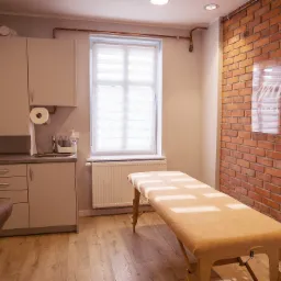 Jasny gabinet z łóżkiem do masażu, ceglaną ścianą z logo, kuchennym aneksem i oknem z żaluzjami.