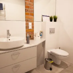 Nowoczesna łazienka z umywalką nablatową, szarą szafką z szufladami, toaletą podwieszaną, koszem na śmieci ze żółtą folią, fragmentem ściany z cegły, plakatem z instrukcją mycia rąk oraz ręcznikiem...