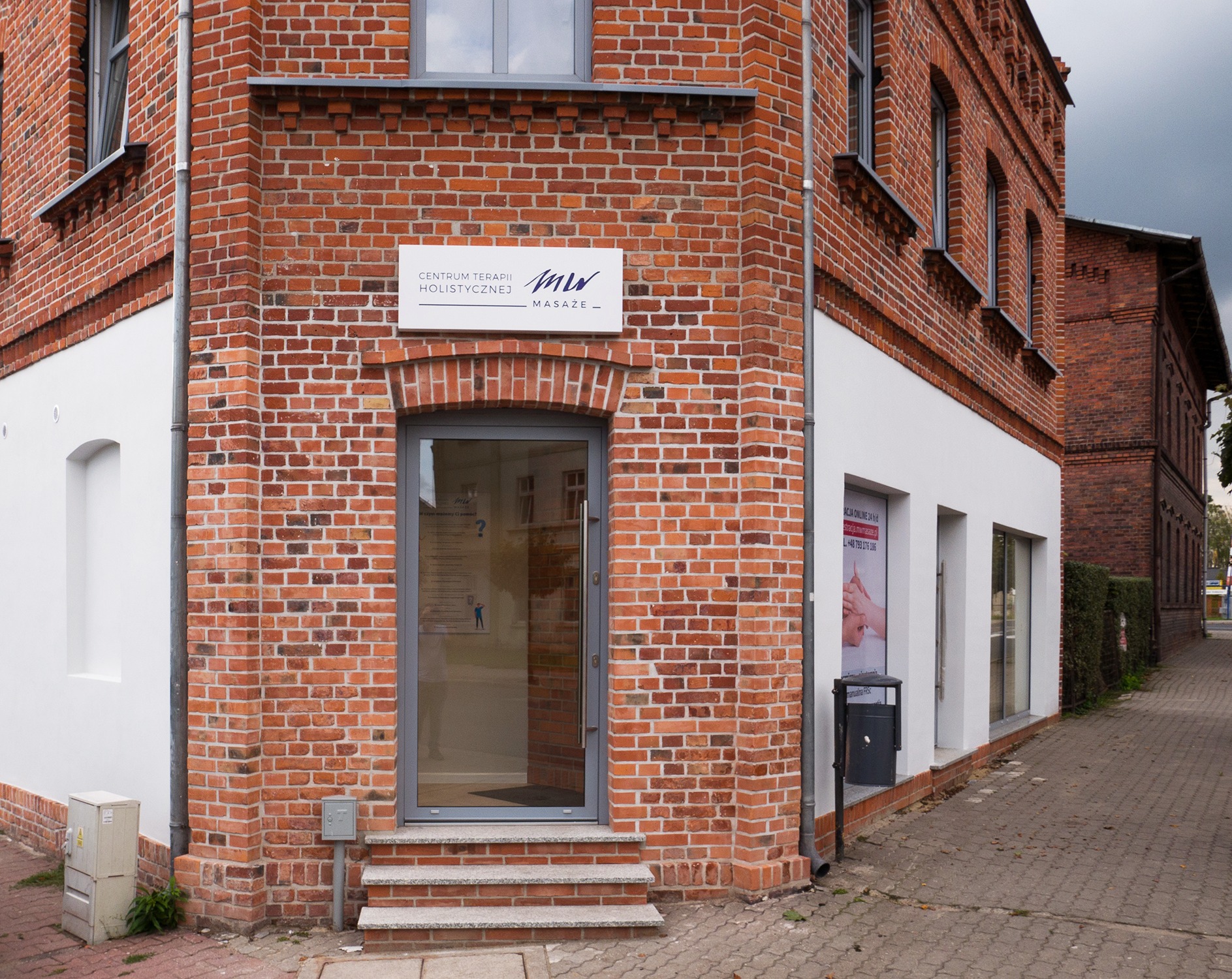 Budynek z czerwonej cegły z szyldem 'Centrum Terapii Holistycznej MW-Masaże' nad wejściem, widoczny fragment chodnika i sąsiedni budynek.