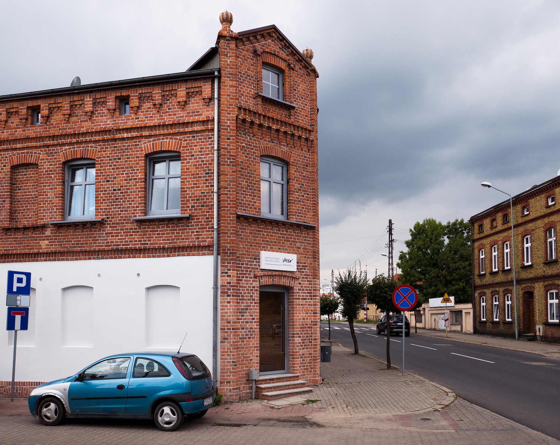 Wejście do Centrum Terapii Holistycznej Masaże w budynku z czerwonej cegły z niebieskim samochodem zaparkowanym obok, widoczne znaki drogowe i ulica.
