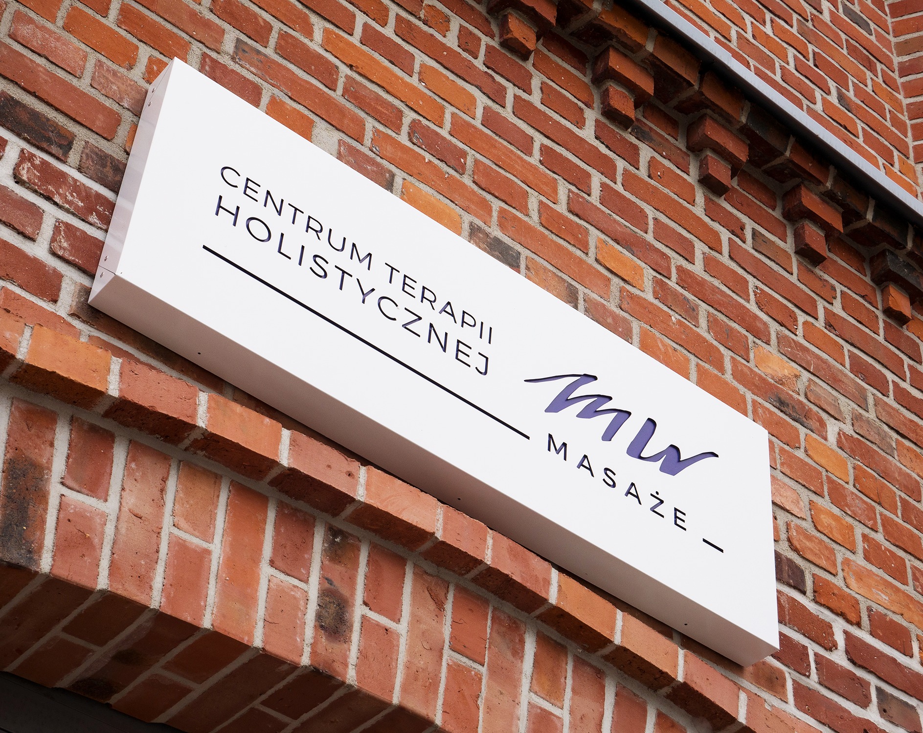 Szyld na ceglanej ścianie z napisem 'Centrum Terapii Holistycznej MW Masaże' w kolorze fioletowym. Minimalistyczny design, nowoczesny font.