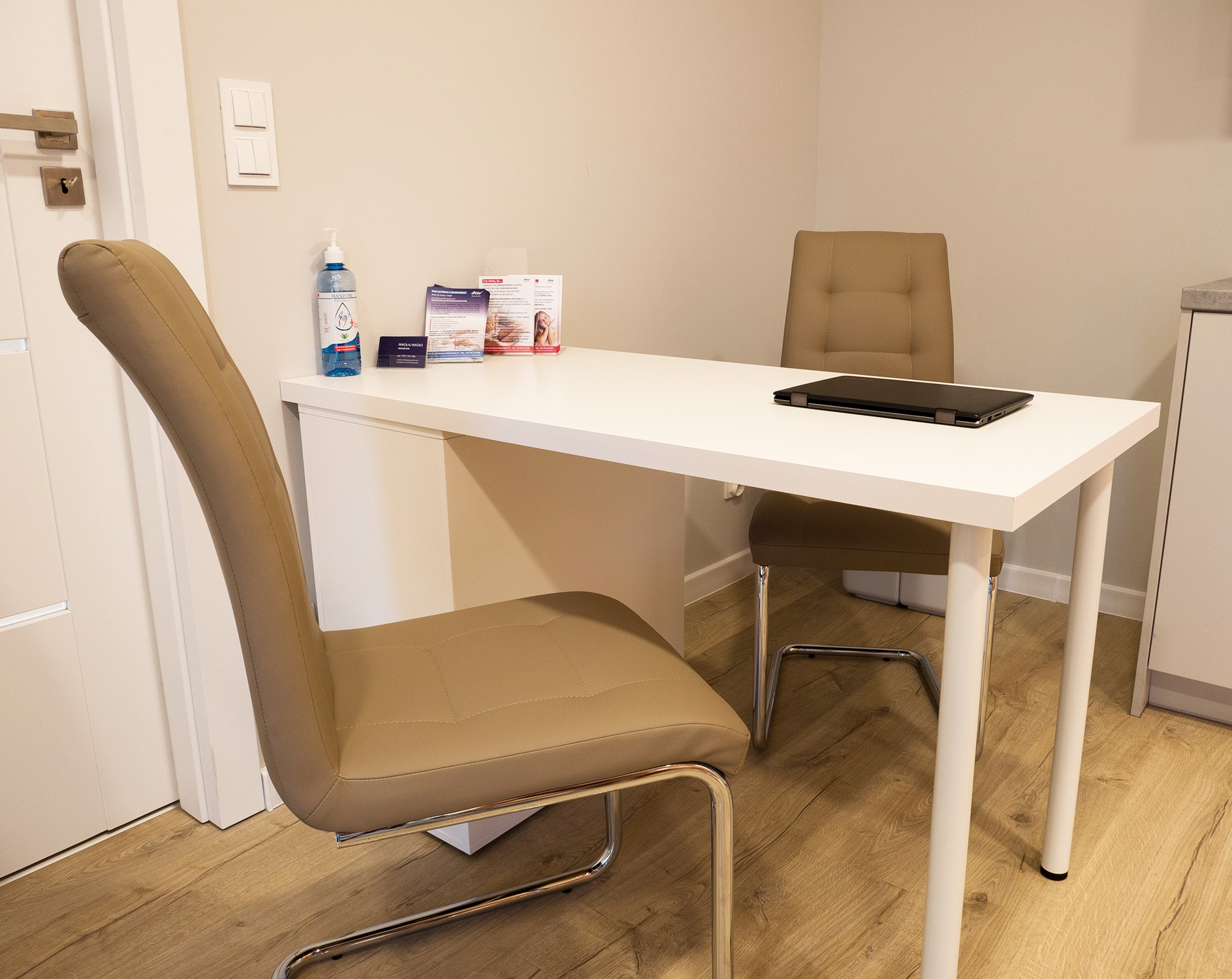 Gabinet: białe biurko z laptopem, ulotki informacyjne, żel antybakteryjny, krzesła w odcieniach beżu, jasne ściany i podłoga.